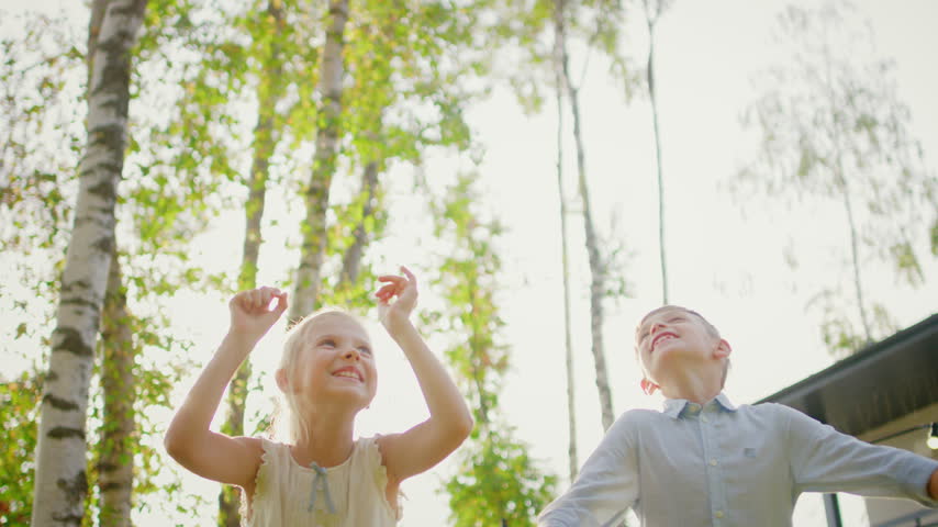 Low Angle Portrait of a Cheerful Little Boy and Girl Laughing and Bouncing High on a Trampoline, Having Fun Together While Their Family Enjoys a Backyard Grill Gathering on a Sunny Summer Day.