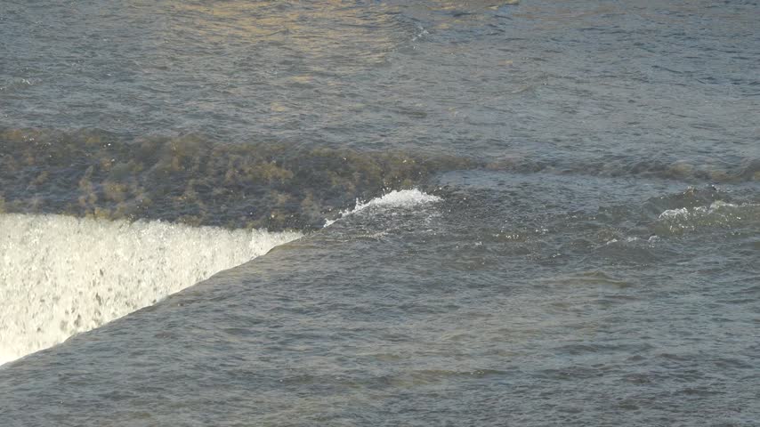 Medium close up slow motion shot of river water cascading off a rubber dam structure, creating strong impact splashes and heavy white foam.
