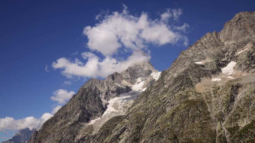 Timelapse of mountains and rapidly moving clouds along the Tour du Mont Blanc, capturing the changing atmosphere of one of the most spectacular trekking regions in the European Alps. Dramatic peaks, alpine ridges, deep valleys, glaciers, and open mountain slopes appear and disappear as clouds drift across the landscape. The accelerated movement of mist, fog, and changing weather creates a dynamic contrast with the rugged terrain, revealing the constantly evolving character of the high alpine environment. Sunlight breaking through the cloud cover can illuminate individual slopes and distant summits, creating shifting patterns of light and shadow across the scenery. The Tour du Mont Blanc passes through France, Italy, and Switzerland, offering exceptional views of the Mont Blanc massif and surrounding mountain ranges. This footage captures the natural beauty, powerful weather, and sense of wilderness associated with this iconic long distance hiking route, making it suitable for travel, adventure, nature, weather, outdoor recreation, and landscape content.