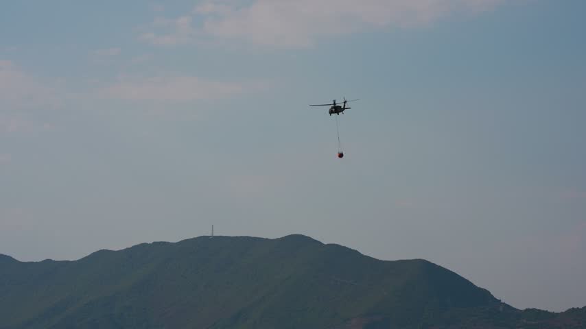 Helicopter with full water bucket ascending over mountain village to fight forest fires