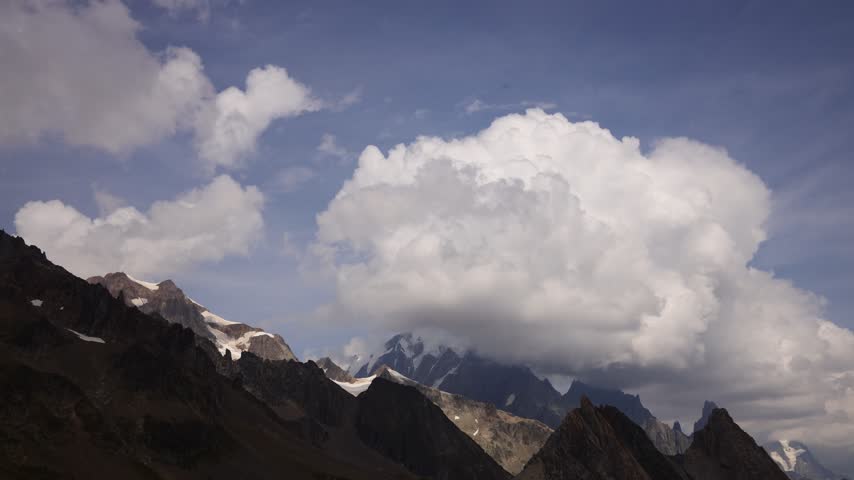 Timelapse of mountains and rapidly moving clouds along the Tour du Mont Blanc, capturing the changing atmosphere of one of the most spectacular trekking regions in the European Alps. Dramatic peaks, alpine ridges, deep valleys, glaciers, and open mountain slopes appear and disappear as clouds drift across the landscape. The accelerated movement of mist, fog, and changing weather creates a dynamic contrast with the rugged terrain, revealing the constantly evolving character of the high alpine environment. Sunlight breaking through the cloud cover can illuminate individual slopes and distant summits, creating shifting patterns of light and shadow across the scenery. The Tour du Mont Blanc passes through France, Italy, and Switzerland, offering exceptional views of the Mont Blanc massif and surrounding mountain ranges. This footage captures the natural beauty, powerful weather, and sense of wilderness associated with this iconic long distance hiking route, making it suitable for travel, adventure, nature, weather, outdoor recreation, and landscape content.
