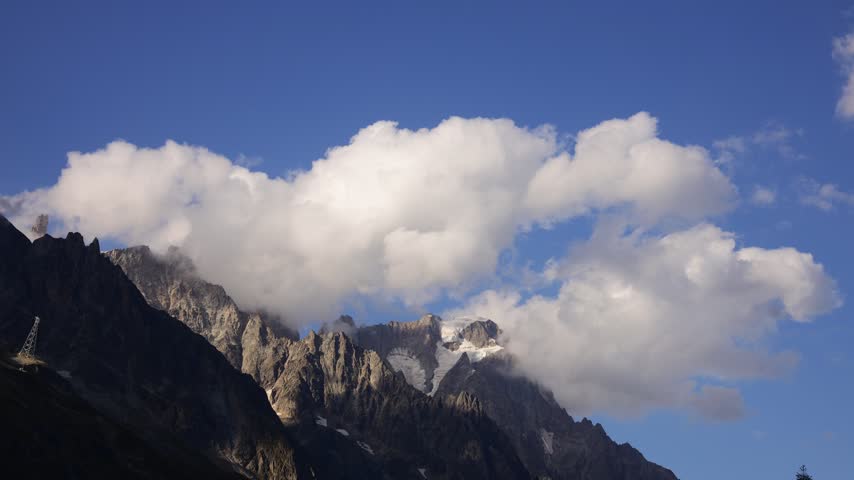 Timelapse of mountains and rapidly moving clouds along the Tour du Mont Blanc, capturing the changing atmosphere of one of the most spectacular trekking regions in the European Alps. Dramatic peaks, alpine ridges, deep valleys, glaciers, and open mountain slopes appear and disappear as clouds drift across the landscape. The accelerated movement of mist, fog, and changing weather creates a dynamic contrast with the rugged terrain, revealing the constantly evolving character of the high alpine environment. Sunlight breaking through the cloud cover can illuminate individual slopes and distant summits, creating shifting patterns of light and shadow across the scenery. The Tour du Mont Blanc passes through France, Italy, and Switzerland, offering exceptional views of the Mont Blanc massif and surrounding mountain ranges. This footage captures the natural beauty, powerful weather, and sense of wilderness associated with this iconic long distance hiking route, making it suitable for travel, adventure, nature, weather, outdoor recreation, and landscape content.