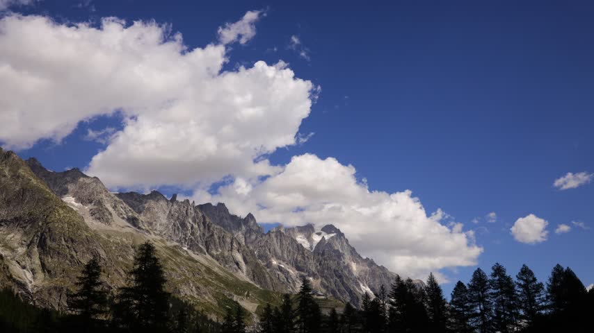 Timelapse of mountains and rapidly moving clouds along the Tour du Mont Blanc, capturing the changing atmosphere of one of the most spectacular trekking regions in the European Alps. Dramatic peaks, alpine ridges, deep valleys, glaciers, and open mountain slopes appear and disappear as clouds drift across the landscape. The accelerated movement of mist, fog, and changing weather creates a dynamic contrast with the rugged terrain, revealing the constantly evolving character of the high alpine environment. Sunlight breaking through the cloud cover can illuminate individual slopes and distant summits, creating shifting patterns of light and shadow across the scenery. The Tour du Mont Blanc passes through France, Italy, and Switzerland, offering exceptional views of the Mont Blanc massif and surrounding mountain ranges. This footage captures the natural beauty, powerful weather, and sense of wilderness associated with this iconic long distance hiking route, making it suitable for travel, adventure, nature, weather, outdoor recreation, and landscape content.