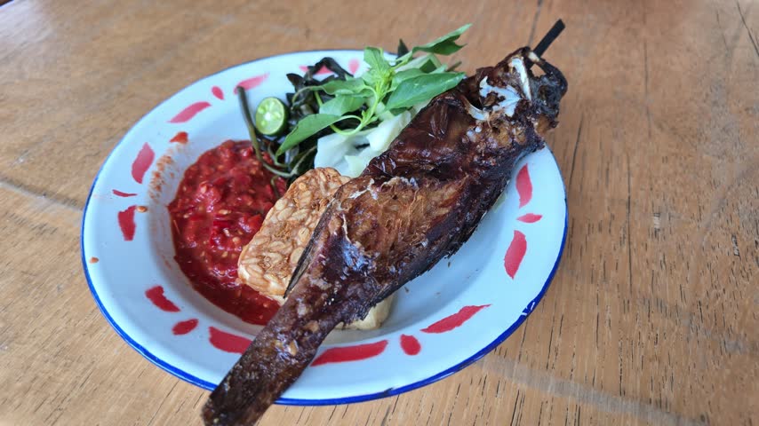 High angle view of whole fried grouper fish on a skewer served on a classic enamel plate with red chili sambal, tempeh, and fresh herbs.
