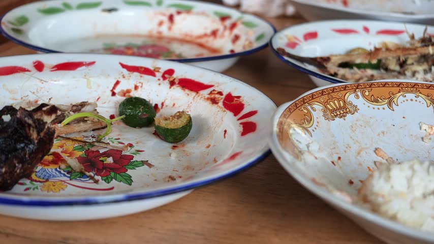 Wide panning shot showing multiple dirty plates, bowls with leftover rice, fish bones, and empty glasses scattered on a restaurant table waiting to be cleaned.
