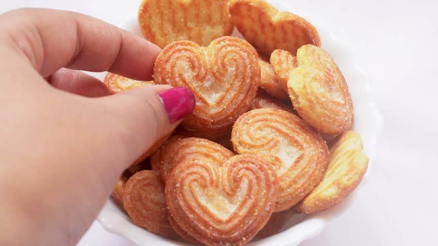Woman hand holding a small heart shape cookies biscuit sprinkled with sugar granules on a white bowl, tea time snack concept.