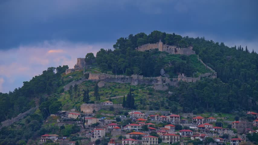 Telephoto shot of the medieval Venetian castle of Nafpaktos perched atop a green hill with traditional red-roofed houses below, framed by dramatic twilight skies in Aetolia-Acarnania, Greece.