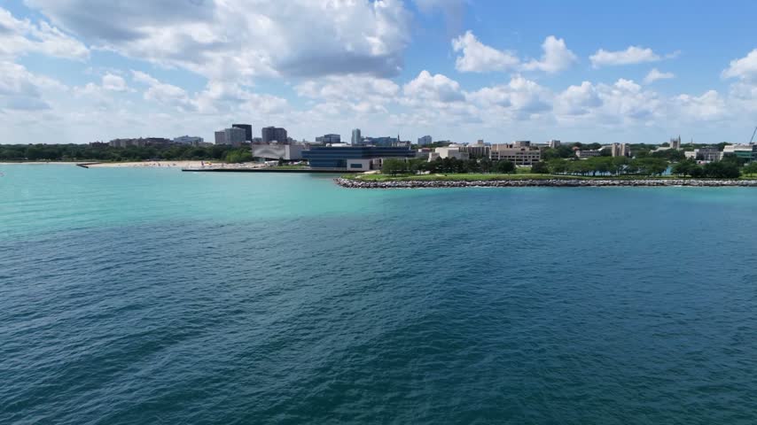 Aerial drone view of Lake Michigan, a sandy Chicago beach, and a lush lakeside park. Blue water, green trees, walking paths, and the urban waterfront create a scenic summer landscape in Chicago, USA