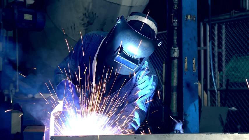 Welder in protective gear works on metal fabrication using welding equipment. Bright sparks fly, highlighting industrial environment with machinery and tools visible.