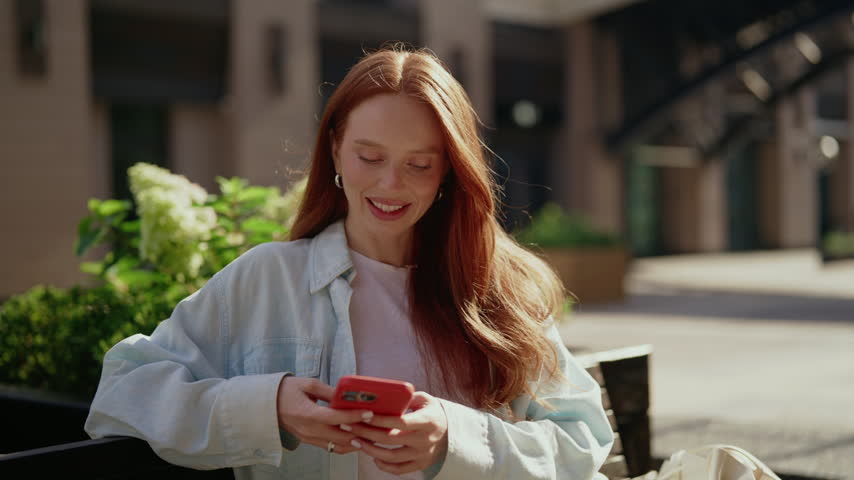 Portrait of beautiful young redhead woman with freckles happily texting on mobile phone. Smiling ginger female sitting on city bench, chatting and browsing social media feed on lovely sunny day.