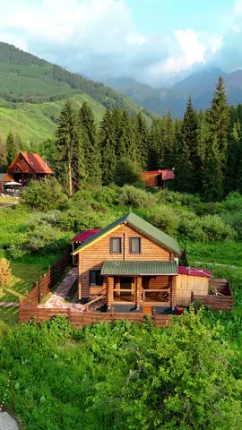 Aerial view of a wooden cabin surrounded by lush vegetation and tall conifer trees in a mountain valley.