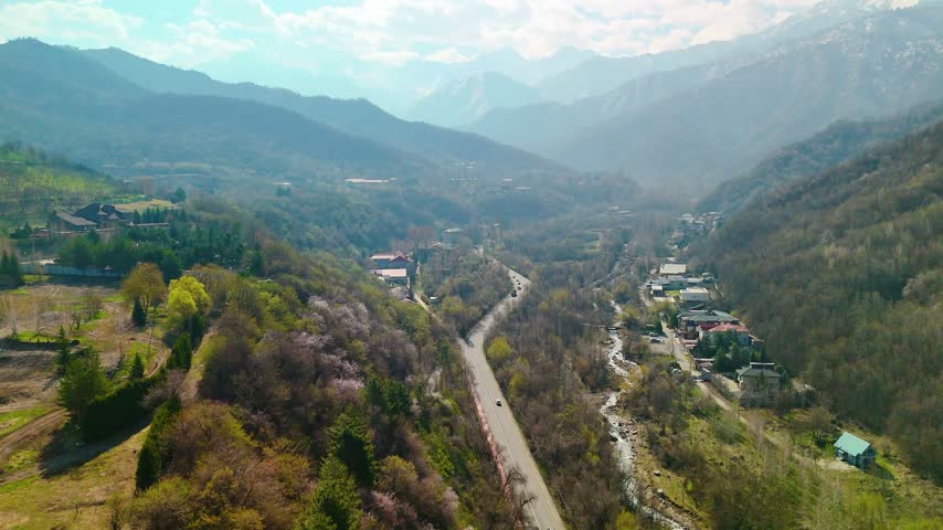 Aerial view of a paved mountain road running beside a rocky stream through a forested valley with scattered houses.