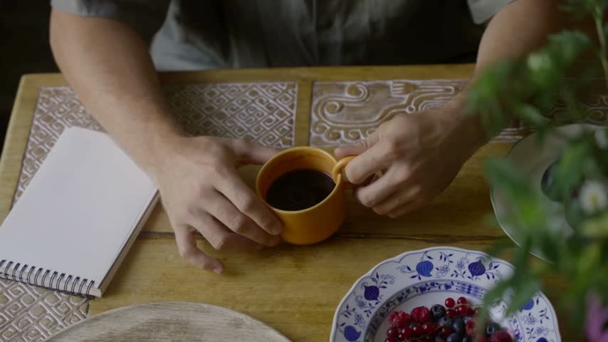 Warm hands gently hold an orange coffee mug, dark liquid inside, on a wooden table. next to fresh berries and blank notebook
