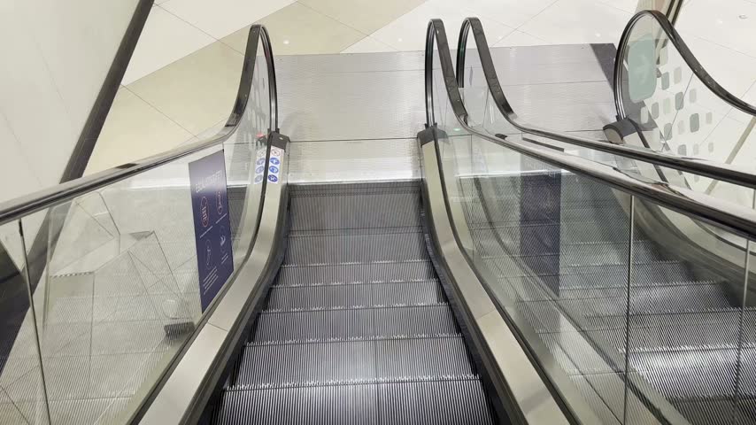Modern escalator steps moving continuously inside shopping mall interior with glass railings and safety signs displayed on metal structures within bright contemporary commercial building hallway