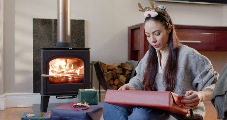 Woman wearing antlers sitting at lit stove, hand passing red gift, checking edges, prepping gifts. Present, wrapping, ribbon, cozy, fireplace, home, festive
