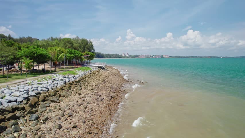 Smooth low-altitude drone flight follows a reinforced tropical shoreline in Malaysia, revealing exposed coastal rocks and gentle waves meeting the coast during low tide.