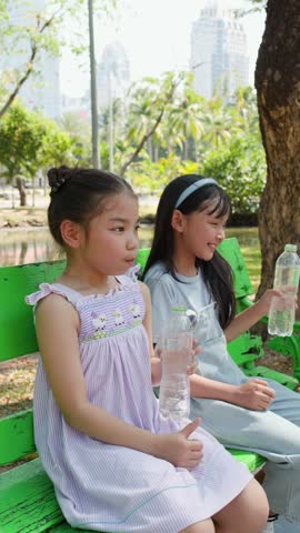 Two young Asian girls laughing on a green bench in Lumpini Park, Bangkok, Thailand.