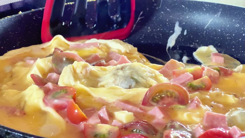 Close-up shot of a red spatula stirring an omelette with diced ham and tomatoes in a skillet. Ideal for breakfast recipes, cookware ads, and culinary videos.