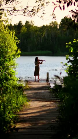 Solitary violinist playing music on a rustic wooden dock by a tranquil forest lake at sunset, surrounded by lush green foliage and calm reflective water