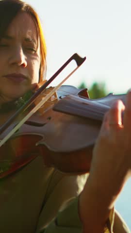 Woman violinist performing outdoors at golden hour, eyes closed in concentration, feeling the emotion of the music against a serene natural background