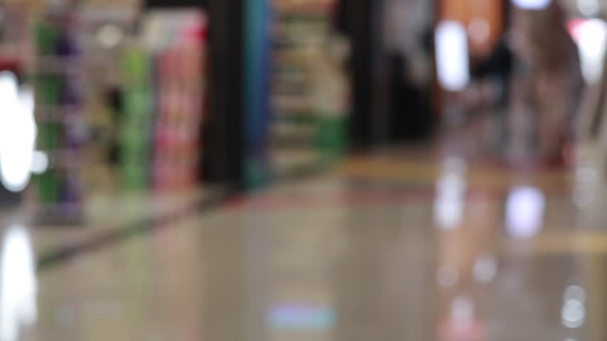 defocused commuters and shoppers walk through a modern mall corridor with reflections on polished floor and colorful storefronts