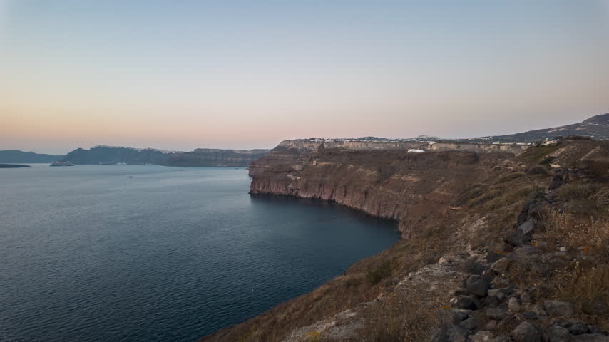 Steep volcanic sea cliffs curve around the quiet Santorini caldera at sunset as warm light fades over the open water.