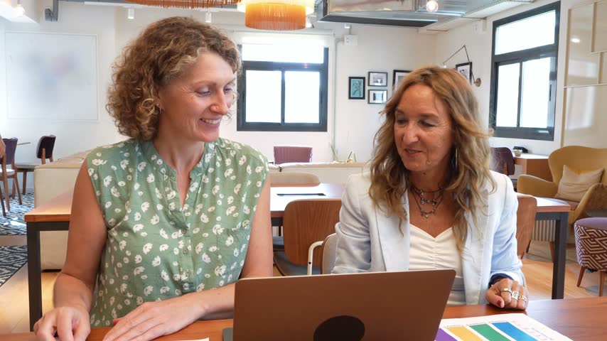 Two smiling mature female coworkers happily discussing business strategy over a laptop and charts in a modern open plan office setting