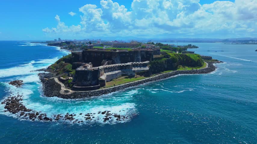 Dramatic high-angle aerial sweep over the historic Spanish fortress San Felipe del Morro with ocean waves crashing against the stone fortifications in Puerto Rico.