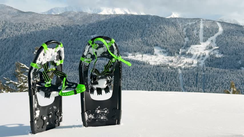 Close up pair of modern black and neon green snowshoes standing in deep snow against scenic mountain range background during sunny winter day.