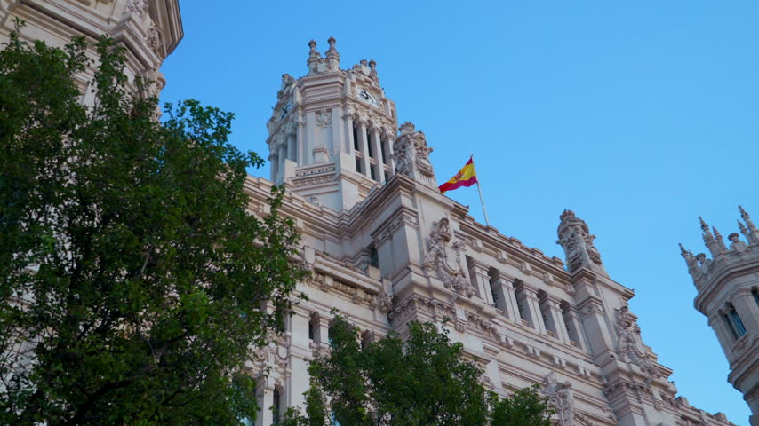 Low angle view of the iconic Cybele Palace in Madrid, Spain, featuring intricate stone carvings and the Spanish flag waving under a clear blue sky.