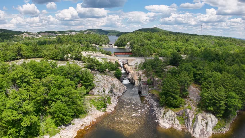 High drone view looks upstream through a rocky forest gorge toward a road bridge crossing above the Whitefish Falls river