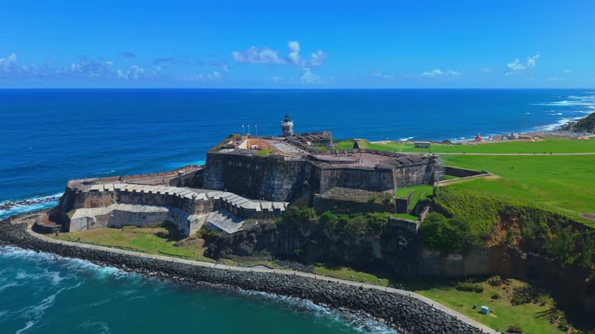 Breathtaking aerial drone footage over Castillo San Felipe del Morro, showing historic stone walls, green lawns, and the Caribbean ocean in Old San Juan, Puerto Rico.