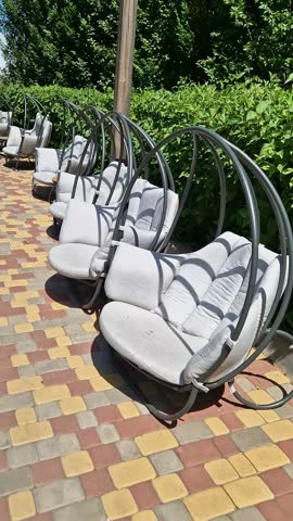 A row of grey hanging pod chairs with soft cushions sits on a colorful paved outdoor patio. Concept of summer relaxation, resort leisure, and peaceful outdoor living environments.