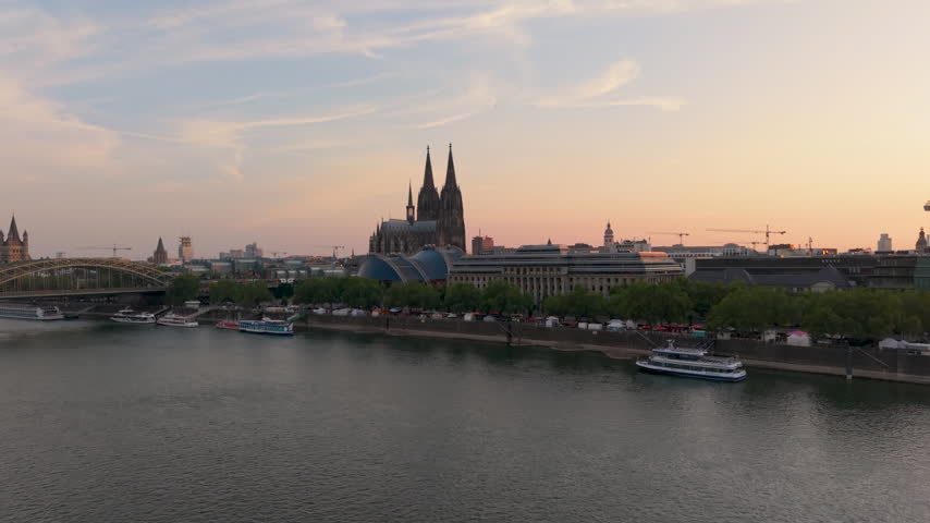 The picture shows Cologne Cathedral in Germany as the sun sets. The cityscape includes buildings, streets, and a river in the background. Many people visit this historic location.