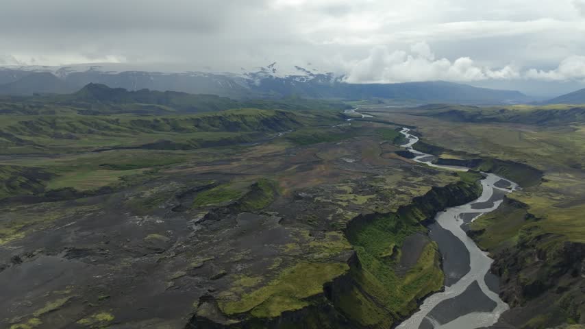 An aerial drone shot pushes in over the vast, rugged landscape of Iceland, revealing the Laugavegur hiking trail winding through valleys, rivers, and volcanic terrain under a cloudy sky.