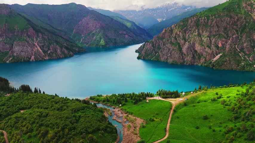 Aerial View of Turquoise Alpine Lake Between Rocky Mountain Cliffs and Green Meadows