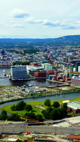 Vertical video. Oslo, Norway. The Oslo Opera House dominates the Bjorvika waterfront, its angular white Carrara marble and granite surfaces creating a striking contemporary form that appears to emerge directly from the dark waters of the Oslofjord. Designed by Snohetta and opened in 2008, the building features a publicly accessible sloping roof that angles down to ground level, forming a broad plaza that invites visitors to walk up and across its surface. The structure is clad in bright white stone that contrasts sharply with the deep blue-grey water and the surrounding modern high-rise developments of the Barcode Project. From this elevated aerial perspective, the opera house sits at the heart of a revitalized harbor district, with boats moored at adjacent piers, construction cranes marking ongoing development, and the dense urban fabric of central Oslo stretching toward forested hills on the horizon. The scene is captured in clear daylight with scattered clouds, revealing the full scope of Norway