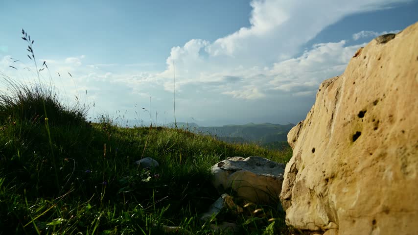 A stationary wide shot of a grassy hill with large rocks and scattered stones in the foreground, looking out toward distant rolling green hills under a cloudy blue sky. The camera is positioned low to the ground with a large rock framing the right side of the frame. Natural daylight illuminates the landscape, with subtle wind movement visible in the grass.