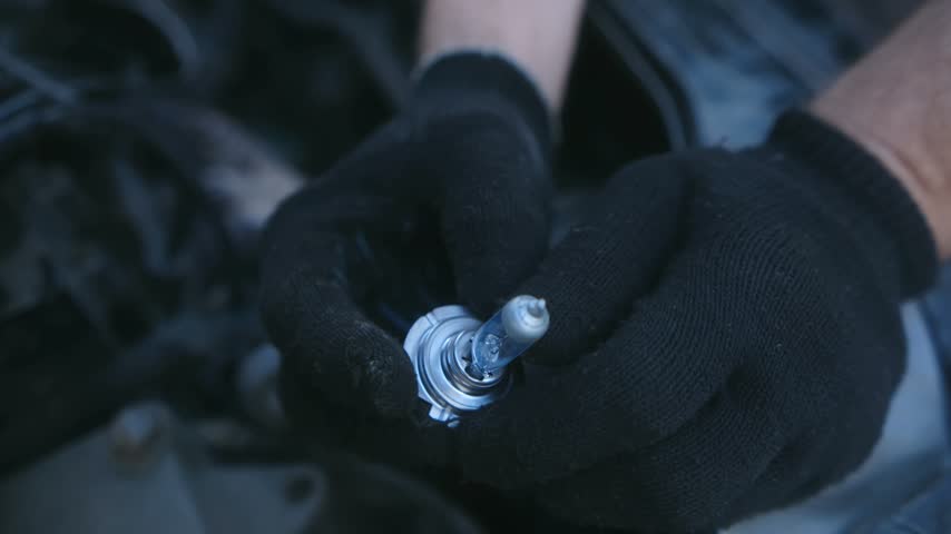 Close up professional auto mechanic wearing black gloves holding new halogen car headlight bulb before installing it in engine bay of modern vehicle during maintenance.