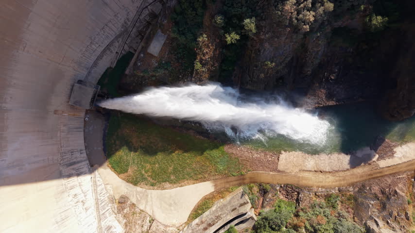 Overhead view of a massive dam spillway releasing powerful white water jet into rocky canyon below, surrounded by green trees and rugged cliff terrain