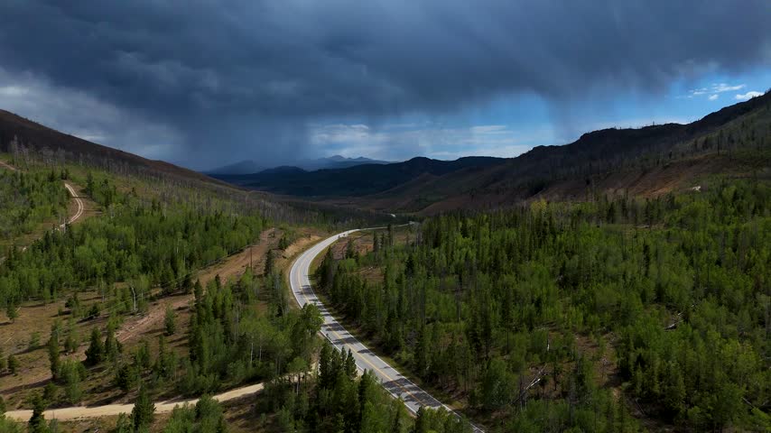 Aerial view of a winding road cutting through a lush green forest in the mountains under a dramatic stormy sky in Granby, Colorado, United States.