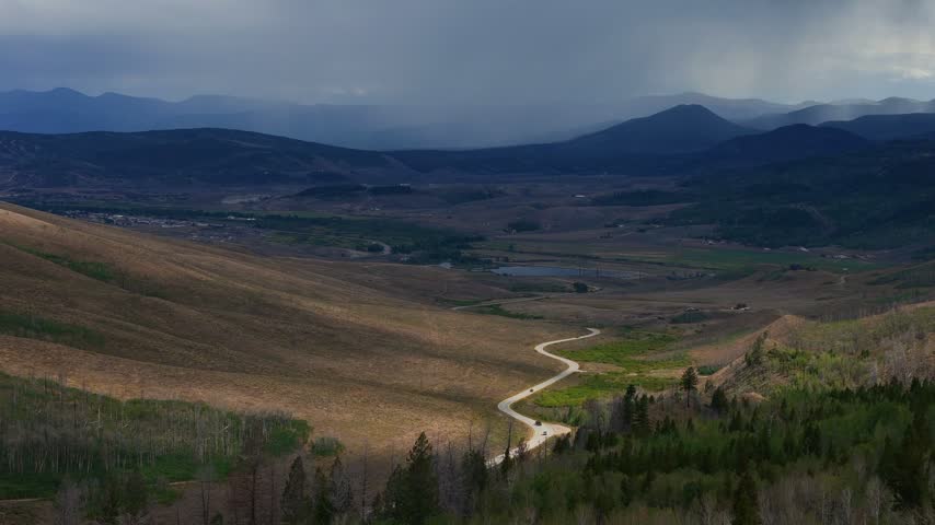 Aerial view of a winding road cutting through a vast valley with rolling hills, pine forests, and distant mountains under a dramatic stormy sky in Granby, Colorado, United States.