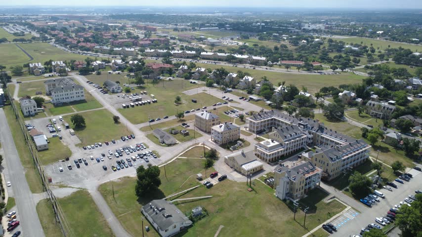 Sprawling masonry compound borders open South Texas airfield near Broadway corridor. Distinct gabled barracks overlook large vehicle lots, access drives, sunny lawns, San Antonio. USA