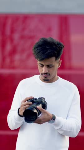 Young moroccan man taking photos with a professional dslr camera. He is adjusting settings, looking through the viewfinder, and shooting. Vertical