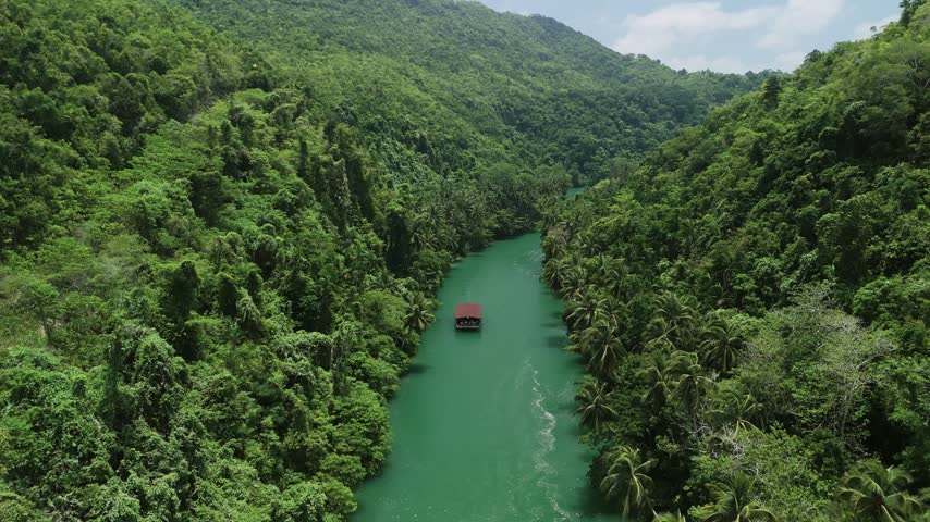 Drone pulls back and ascends as a Loboc River cruise boat travels along the green river between forested hills in Bohol