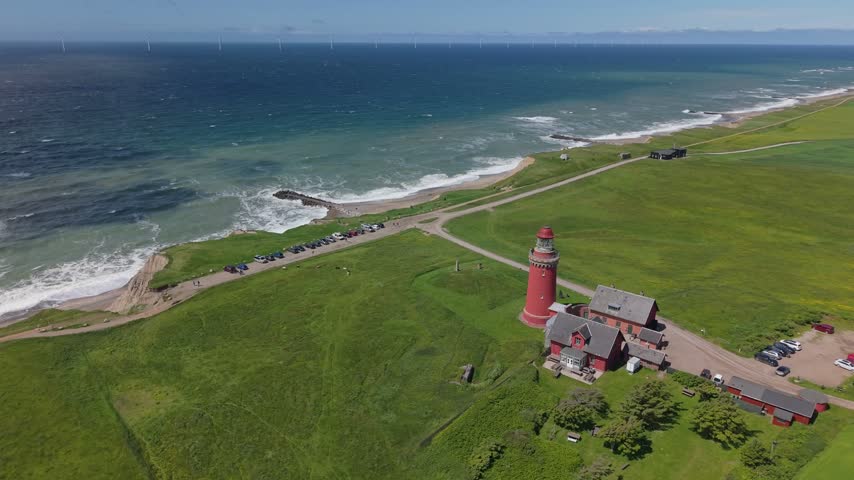 Aerial orbit around Bovbjerg Lighthouse, revealing lush green meadows, rugged coastal cliffs, and the waves of the North Sea along Denmark