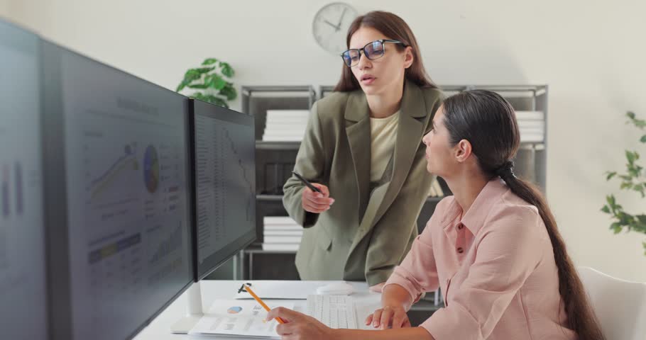 Women analysts discuss financial data analysis at computer. They review charts and KPIs on dual monitors as one colleague guides another. Image conveys leadership coaching and data teamwork.