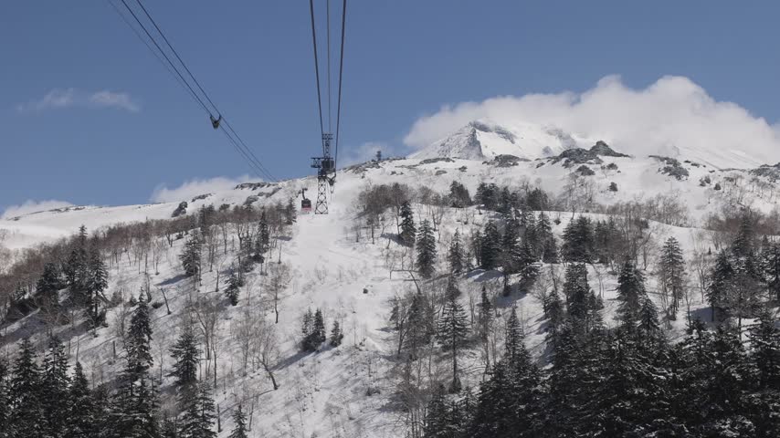 View of the Asahidake Ropeway cable car ascending over snow-covered slopes and evergreen forests in Higashikawa, Hokkaido, Japan.