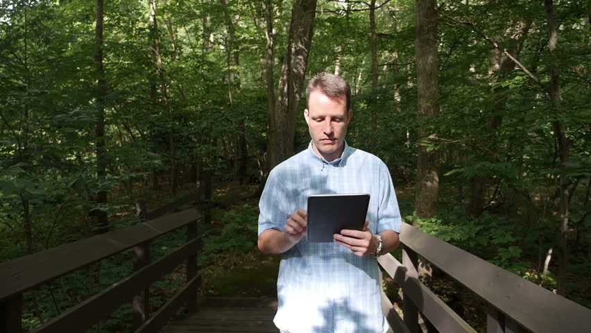 A man with a tablet PC walks through the woods looking at plant life.