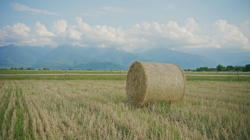 Hay bale sits in a field with mountains in the background under a cloudy sky during daytime in a rural area